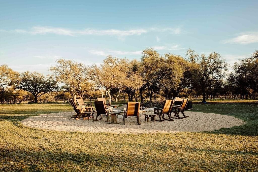 An outdoor seating area with a circular arrangement of wooden chairs and tables, surrounded by trees.