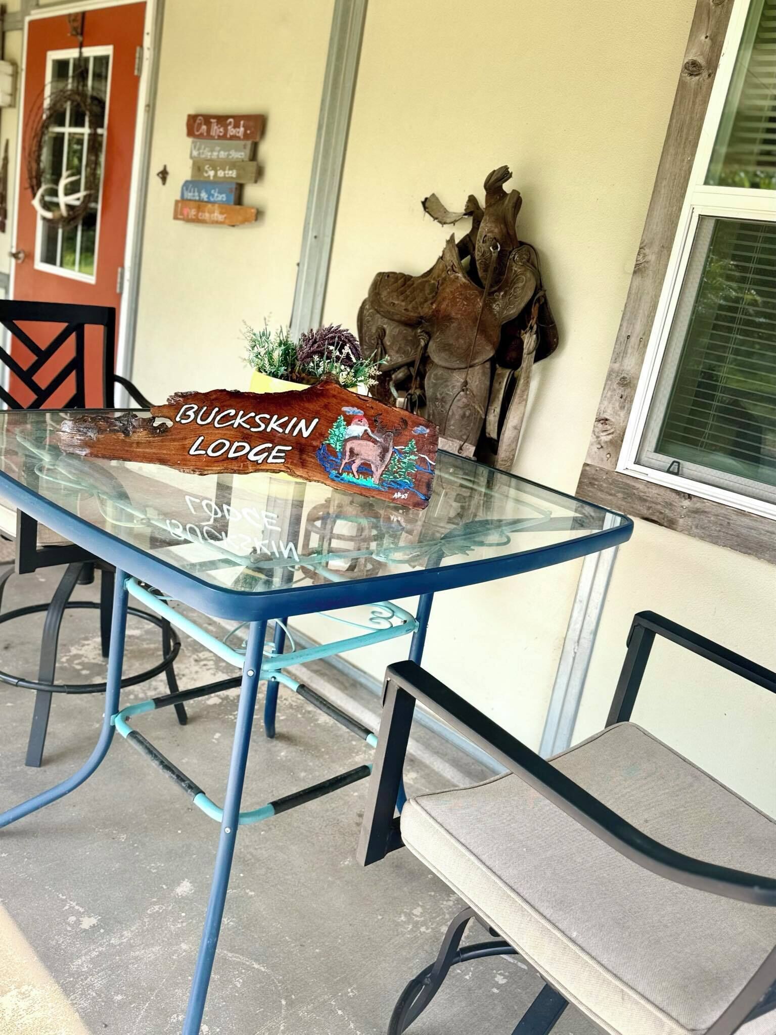 Une table avec un plateau en verre et des pieds en métal, une enseigne en bois portant l’inscription « BUCKSKIN LODGE », et une sculpture de cerf accrochée au mur.