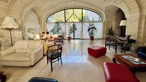 A spacious living room with a high arched ceiling, a large window, and a mix of seating including a sofa, chairs, and a red ottoman. - Albergo Masseria Gattamora (Uggiano la Chiesa)