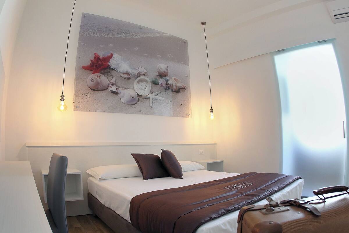 A modern hotel room with a large bed, a desk, and a chair. There is a painting on the wall and a suitcase on the bed.