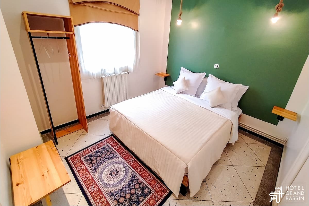 Hotellihuone, jossa on sänky, ikkuna verhoilla, patteri, puinen yöpöytä, riippuvalaisin ja vihreä tehosteseinä.