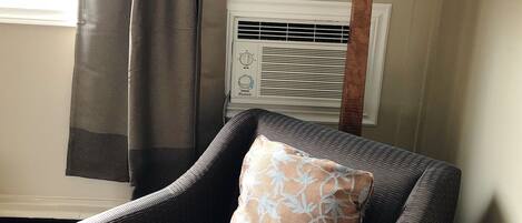 A chair with a patterned cushion, a lamp, and an air conditioning unit in a room with a window and curtains.