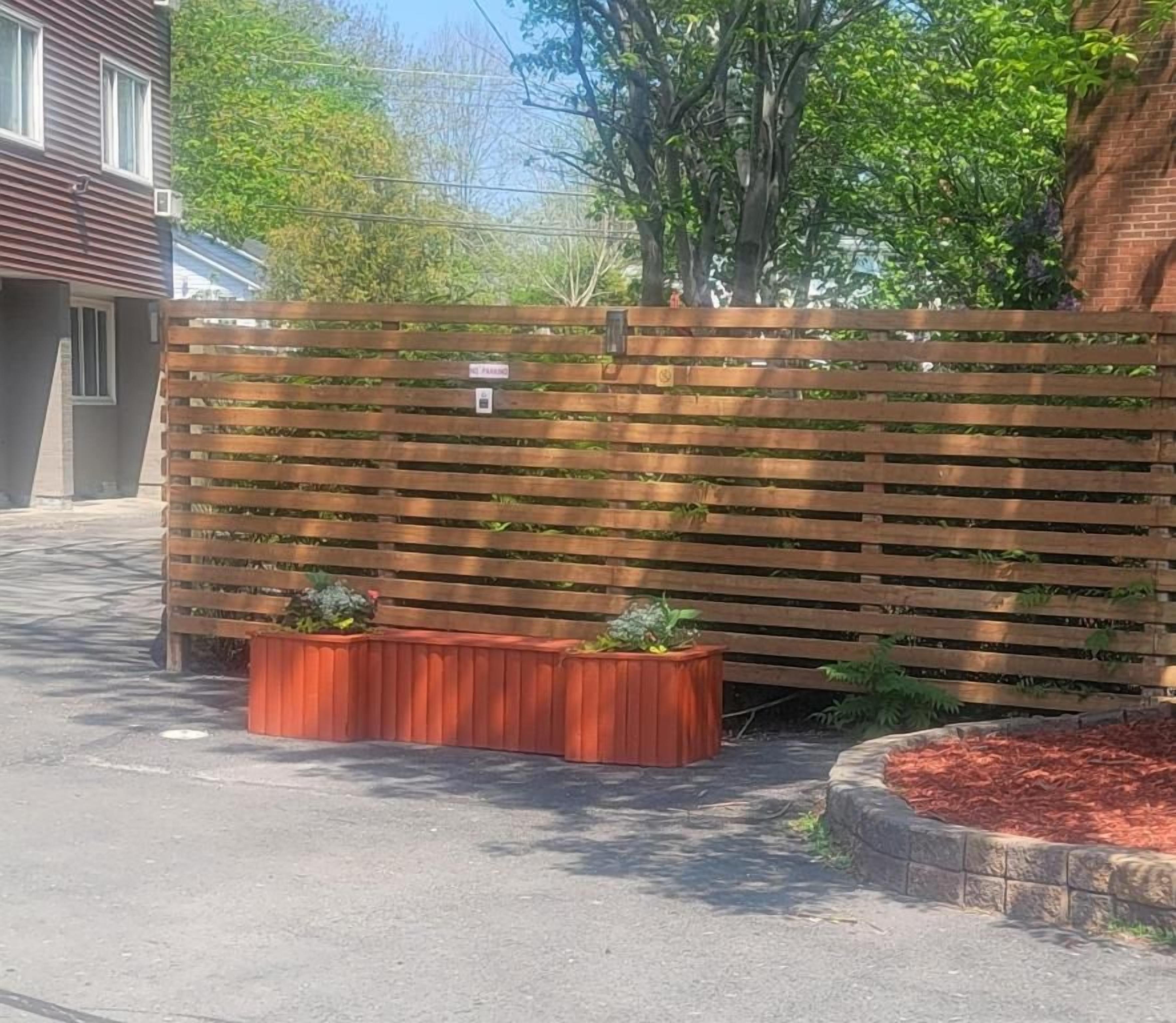 A wooden fence with a small sign, a paved area, and a brick planter with plants.