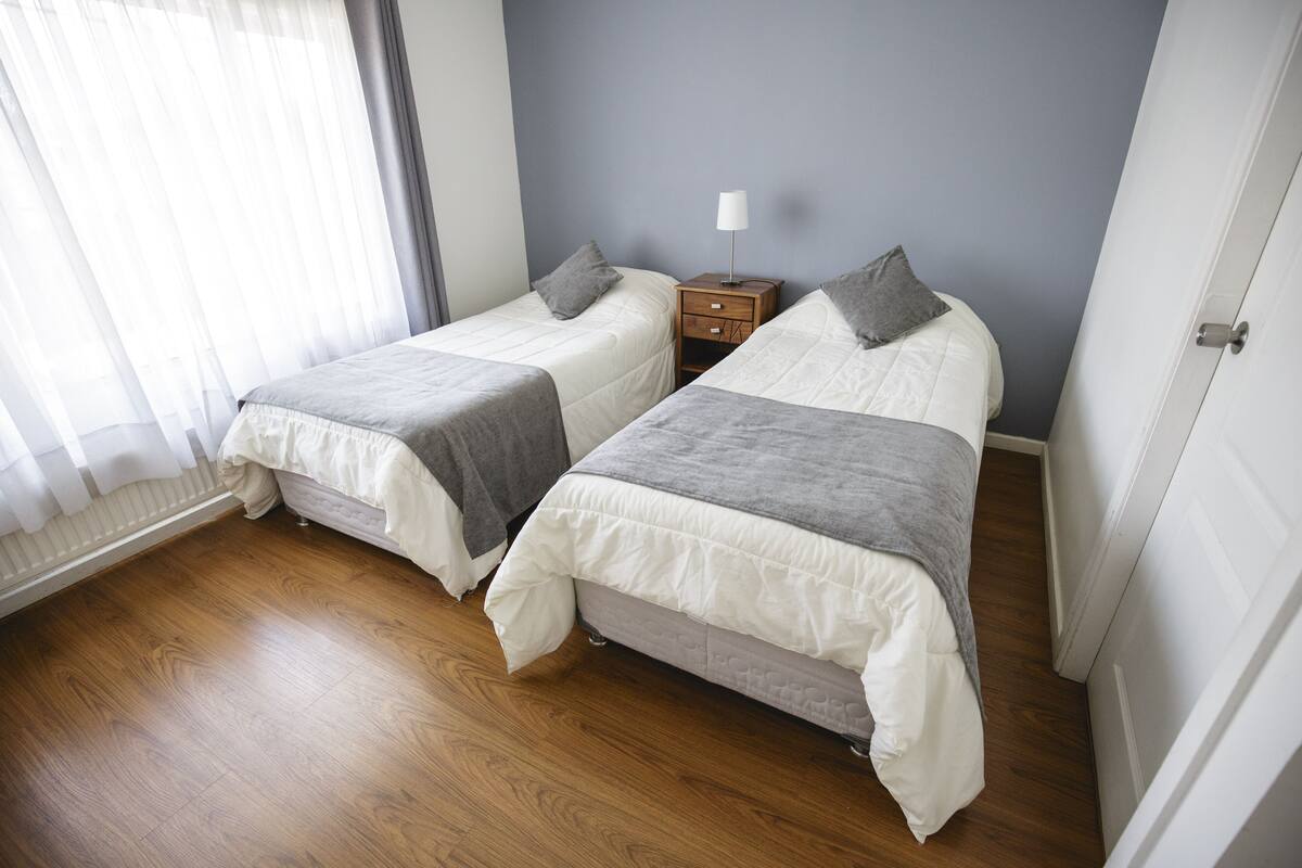 A bedroom with two beds, a nightstand, and a window with sheer curtains.