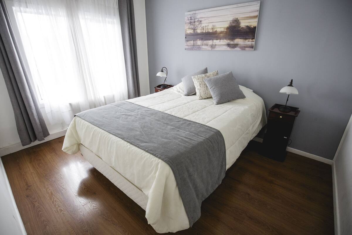 A neatly made bed with a gray and white duvet, two bedside tables with lamps, a painting on the wall, and sheer curtains on the window.