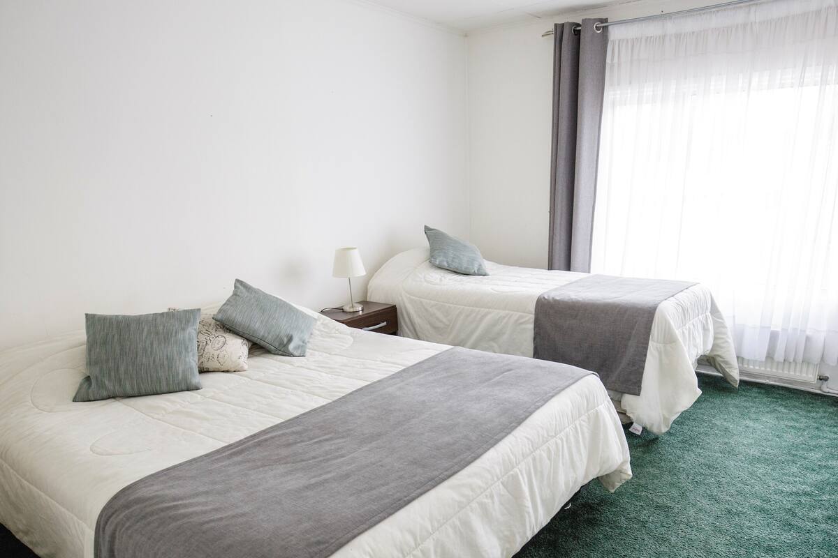 A hotel room with two beds, a nightstand, a lamp, and a window with curtains.