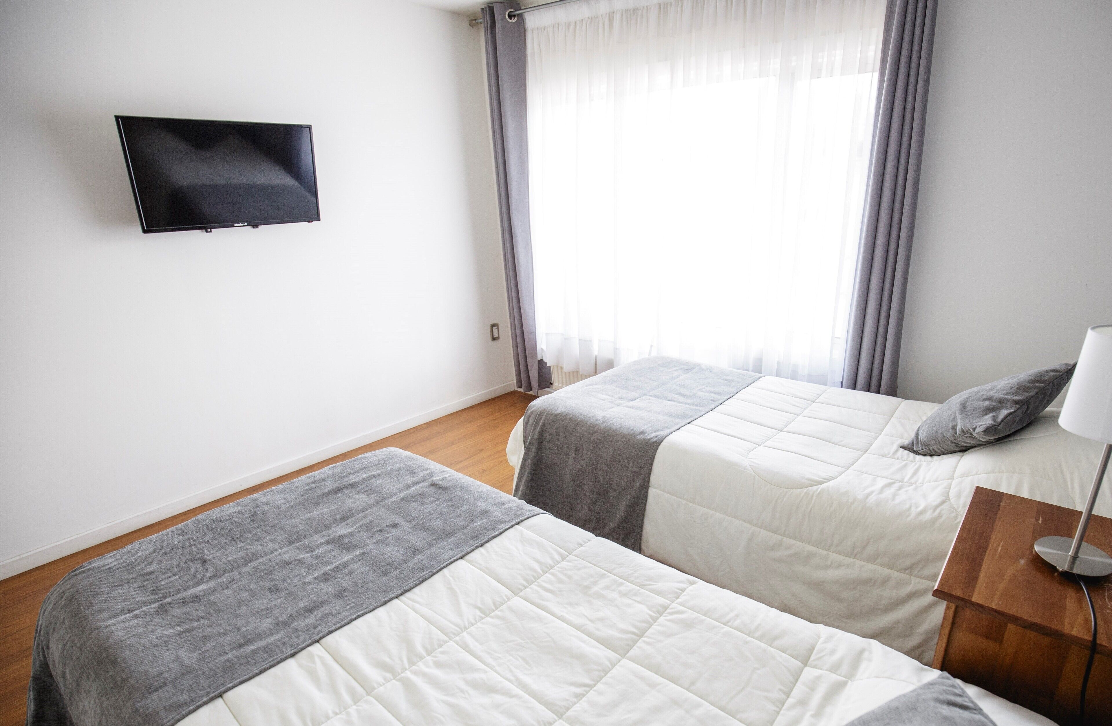 Habitación de hotel con dos camas, un televisor de pantalla plana montado en la pared, una mesita de noche con lámpara y una ventana con cortinas.