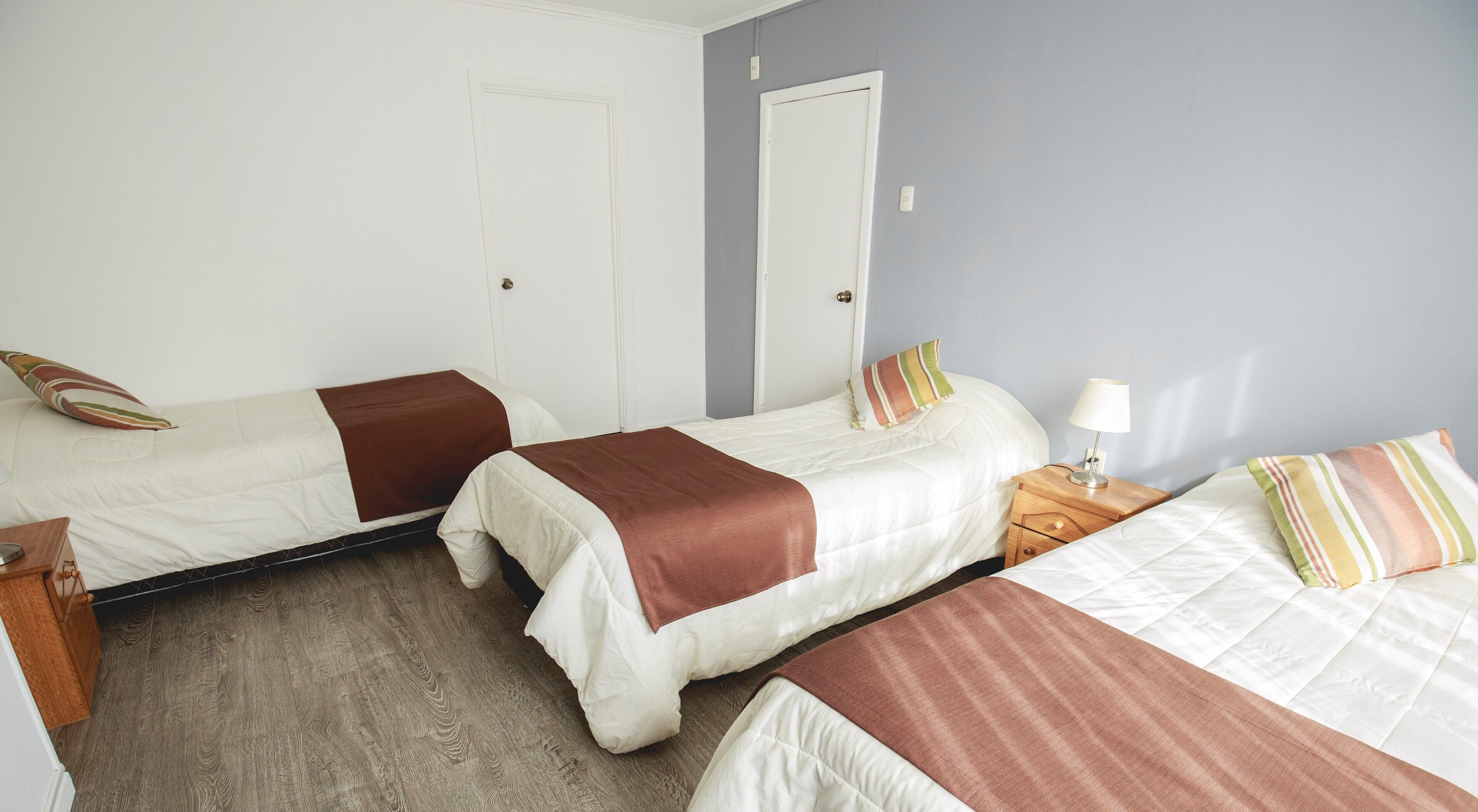A room with three beds, each with a brown blanket. There is a wooden nightstand with a lamp and a phone on it. The room has white walls and a wooden floor.