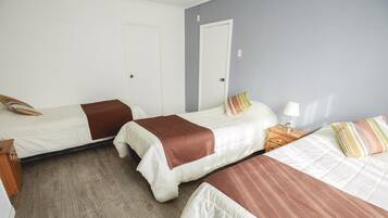 Ein Zimmer mit drei Betten, jeweils mit einer braunen Decke. Ein Holznachttisch mit einer Lampe und einem Telefon steht daneben. Die Wände sind weiß und der Boden ist aus Holz.