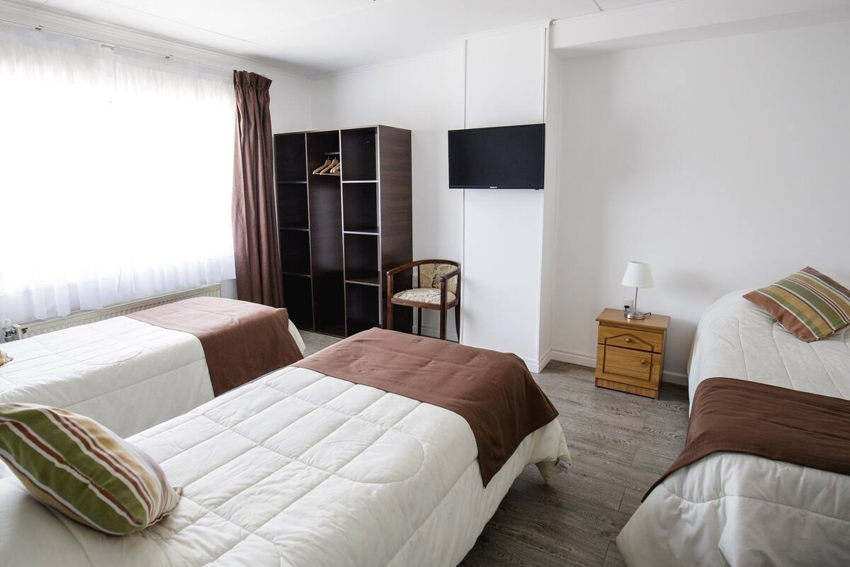A hotel room with two beds, a wardrobe, a chair, a TV, and a bedside table with a lamp.