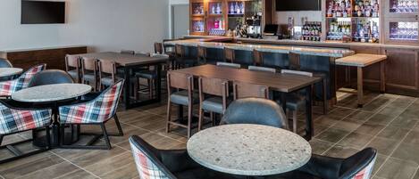 Eine Bar mit verschiedenen Sitzgelegenheiten, darunter runde Tische und Barhocker, sowie einer Theke mit einer Auswahl an Flaschen und Gläsern.