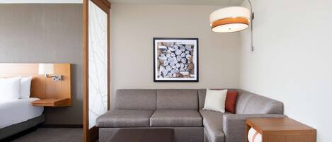 A hotel room with a bed, a gray sofa, a wooden bench, a wall-mounted lamp, and a framed picture.