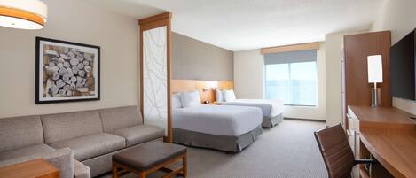 A hotel room with a sofa, two beds, a desk, a chair, a lamp, and a framed picture on the wall.