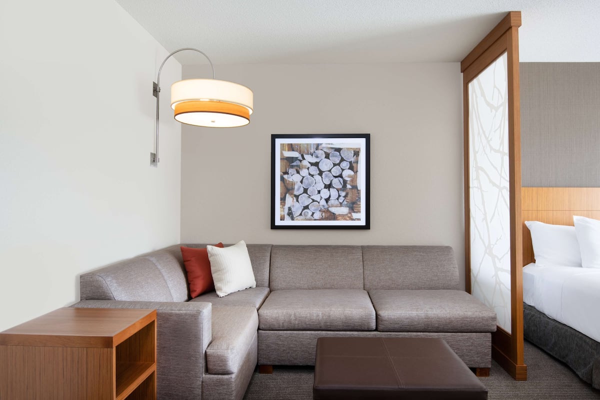 A modern hotel room with a grey sofa, a wooden side table, a framed artwork on the wall, and a bed with white linens.