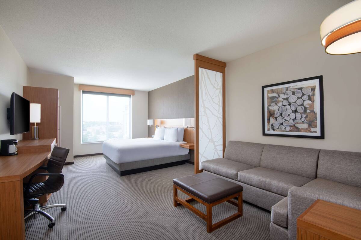 A hotel room with a bed, a sofa, a desk with chairs, a television, and a framed picture on the wall.
