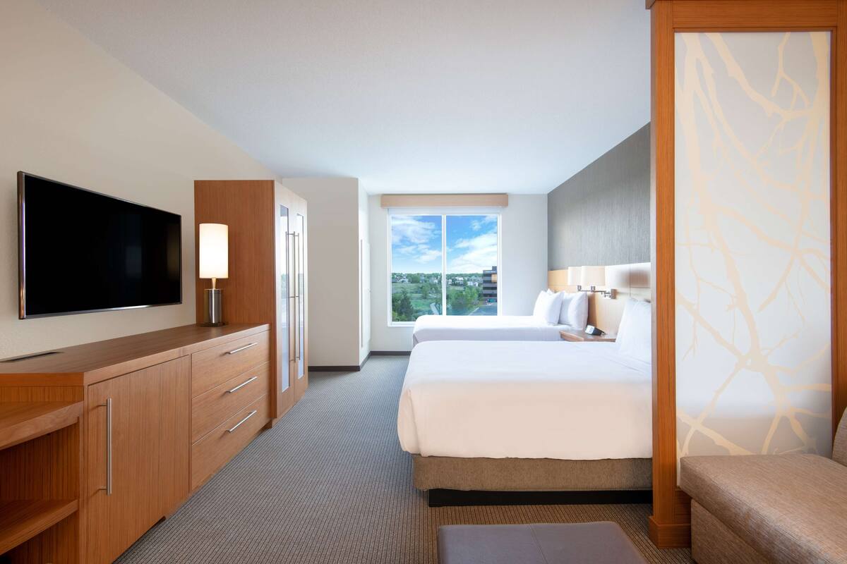 A hotel room with a large flat-screen TV, a bed with white linens, a wooden dresser, a bedside table with a lamp, and a window with a view of the city.