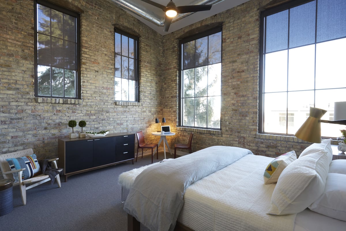 Ein Schlafzimmer mit einem großen Bett, einem Schreibtisch und einem Stuhl. Eine Ziegelwand und große Fenster sind zu sehen.