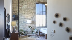 A room with a brick wall, a large window, a bed, two chairs, a small table, a lamp, a wall-mounted TV, and a bookshelf. - Kinn Guesthouse Bay View (Milwaukee)