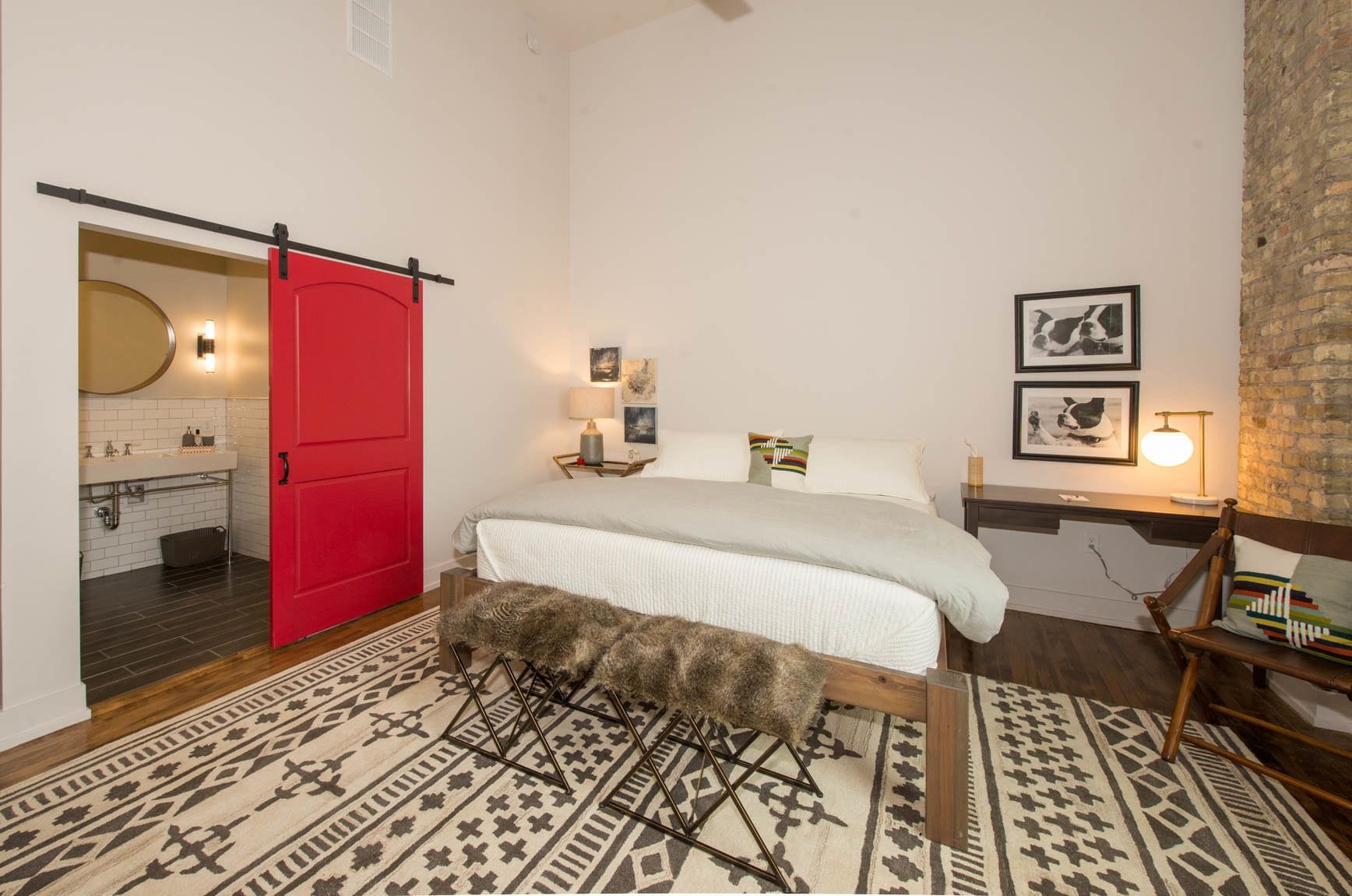 Ein Schlafzimmer mit roter Tür, einem Bett mit weißer Bettdecke, einem gemusterten Teppich, einer hölzernen Bank und einer Ziegelwand.