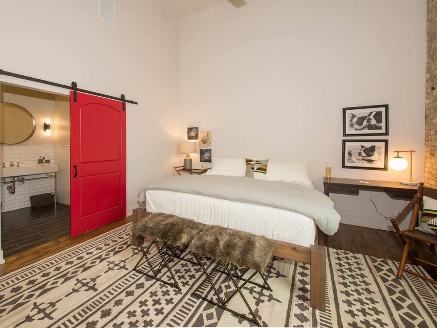 A bedroom with a red door, a bed with a white comforter, a patterned rug, a wooden bench, and a brick wall.