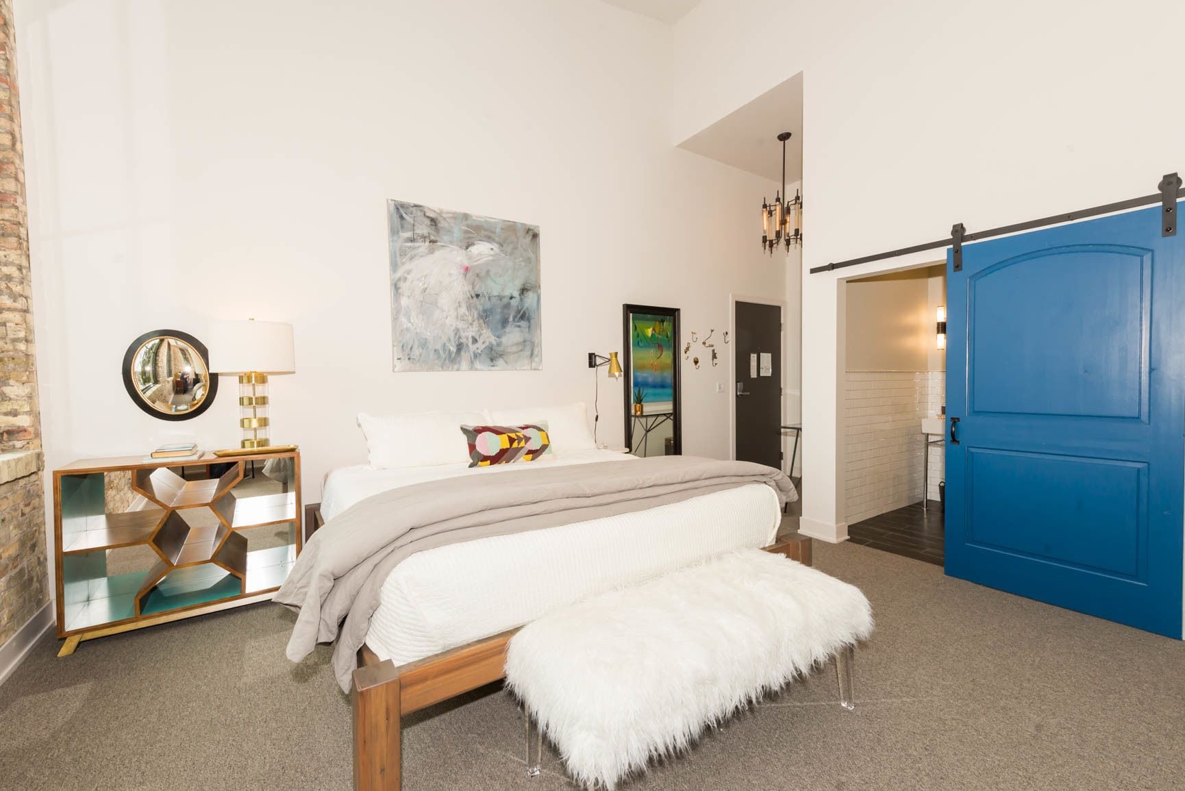Un dormitorio con cama, espejo, pintura, puerta azul y una pequeña banca blanca.