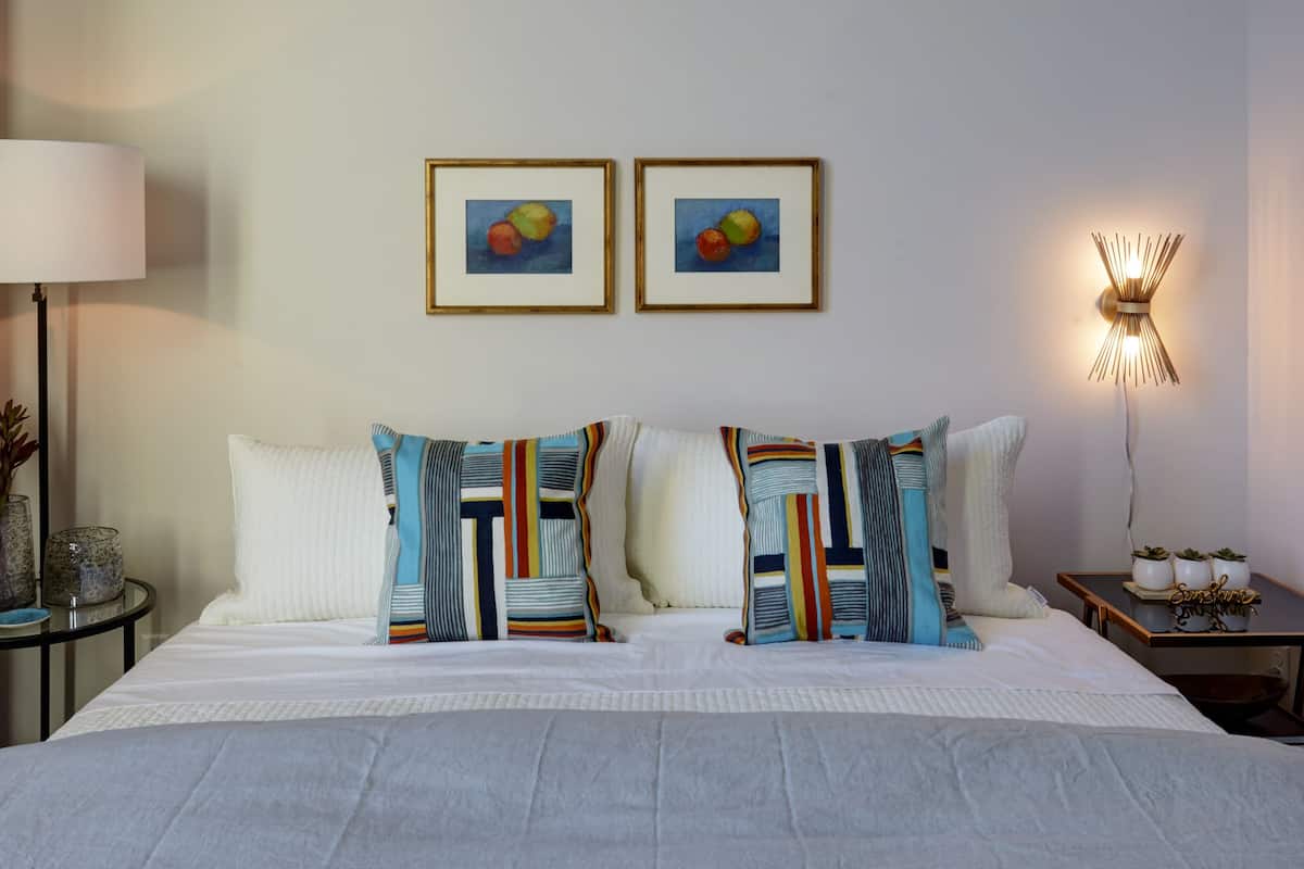 Ein Hotelzimmer mit einem großen Bett, zwei gerahmten Früchtebildern an der Wand, einer Nachttischlampe und einem kleinen Tisch mit einer Pflanze und einem dekorativen Gegenstand.