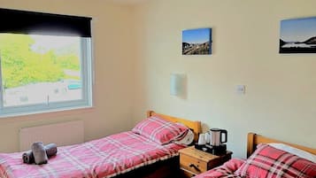 Una habitación con dos camas de madera, cada una con colcha y almohadas a cuadros rojos y blancos. Hay una ventana que da a vistas de vegetación, una lámpara en el techo y dos marcos de cuadros en la pared.