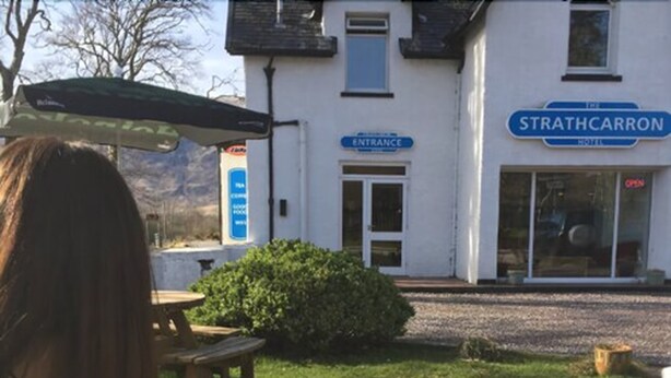 A person sitting at an outdoor table with a drink and a menu, in front of a building labeled "STRATHCARRON HOTEL" with an entrance sign. - Strathcarron Hotel (Strathcarron)