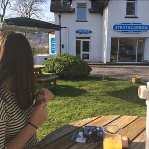 A person sitting at an outdoor table with a drink and a menu, in front of a building labeled "STRATHCARRON HOTEL" with an entrance sign. - Strathcarron Hotel (Strathcarron)