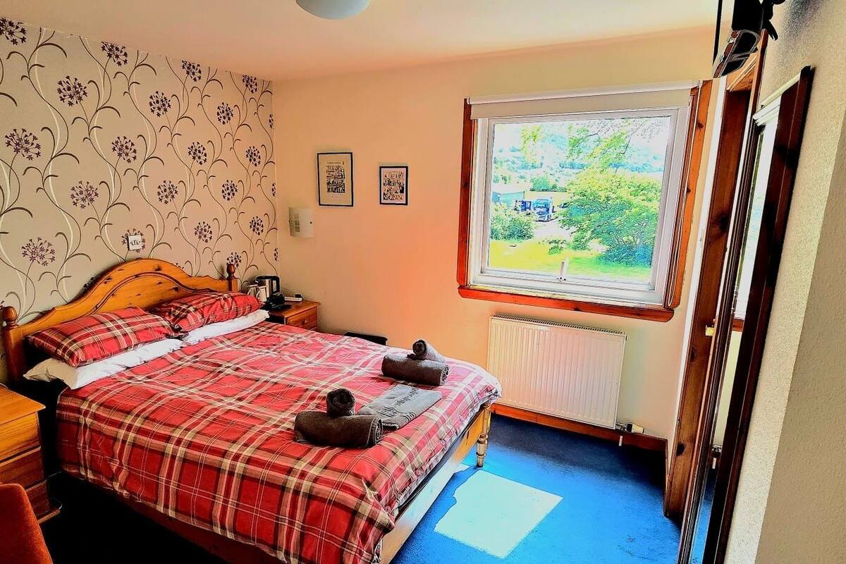 Quarto com cama, mesa de cabeceira, janela com vista para área verde e radiador.
