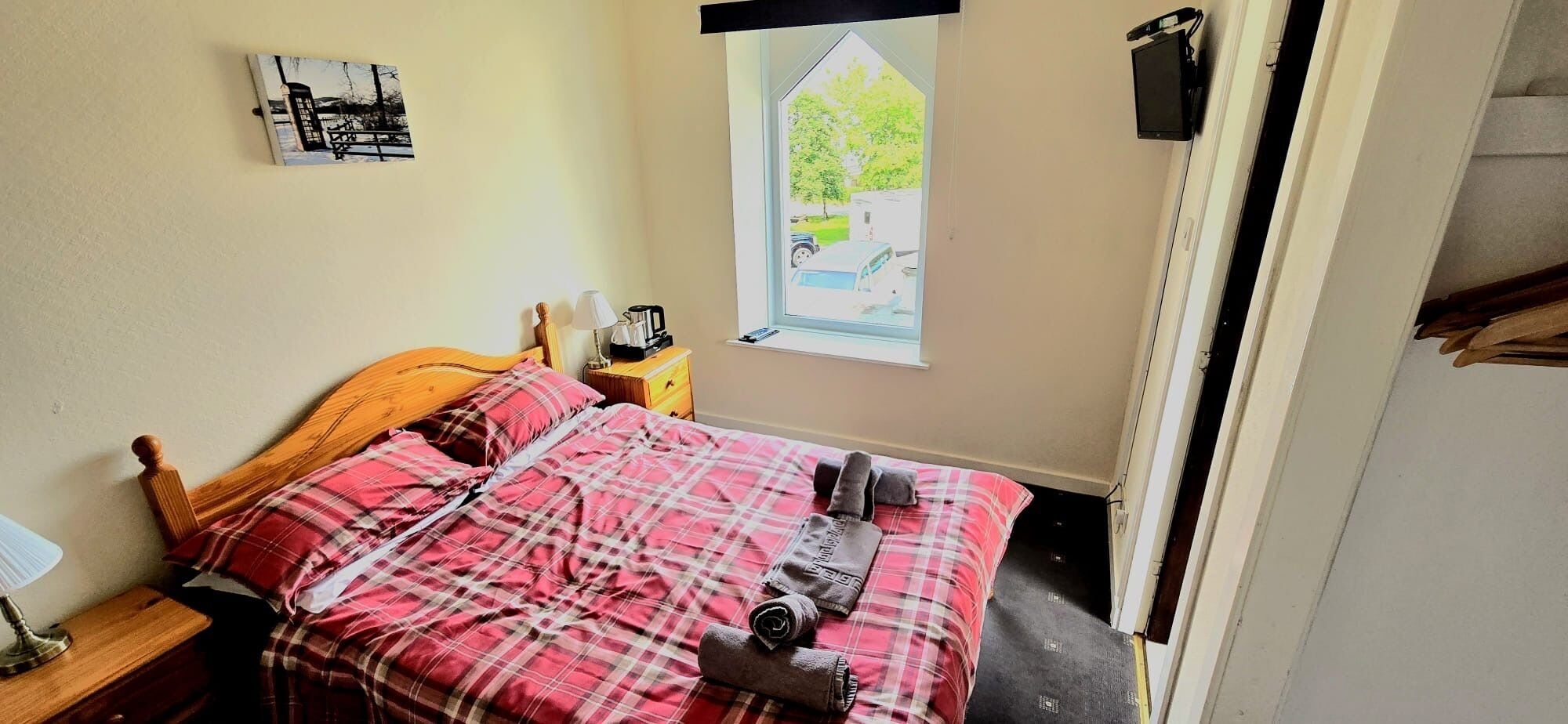 A bedroom with a bed, a nightstand, a lamp, a window with a view of a tree and a car, and a wall-mounted TV.