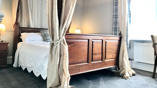 Una cama con dosel y cuatro columnas, con tablero de madera, ropa de cama blanca y una lámpara de noche.