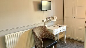 Una habitación con un radiador, un sillón estampado, un escritorio blanco con cajones, un televisor montado en la pared y una puerta.