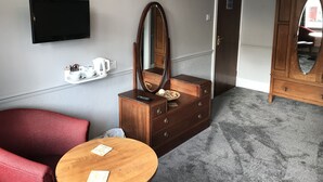 Habitación de hotel con televisión de pantalla plana, cómoda de madera con espejo, una silla roja, una mesita redonda y piso alfombrado.