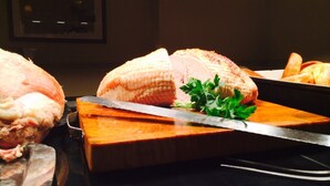 Un pavo entero asado sobre una tabla de cortar con un cuchillo para trinchar, rodeado por una mesa de madera y una encimera negra.
