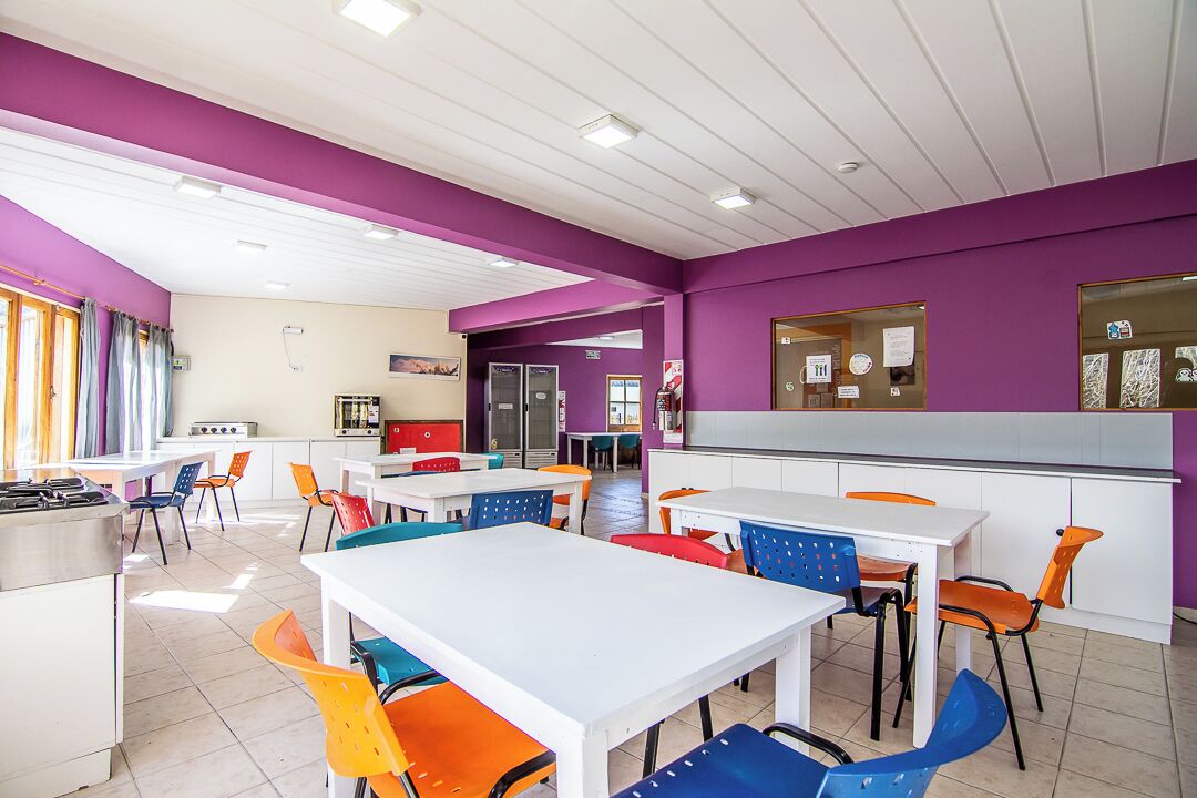 Uma área de refeições com mesas brancas e cadeiras coloridas, paredes roxas e uma cozinha ao fundo.