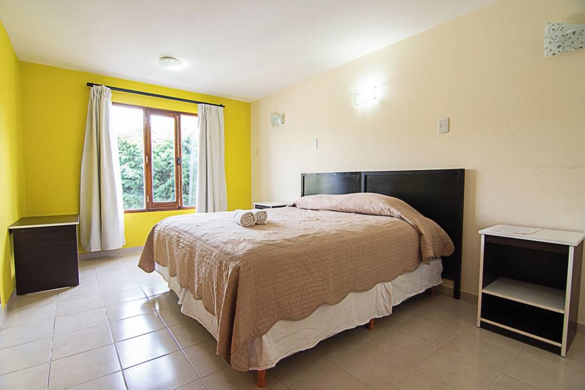 Ein Schlafzimmer mit einem Bett, einem Fenster mit Vorhängen, einem Nachttisch und einem Beistelltisch.
