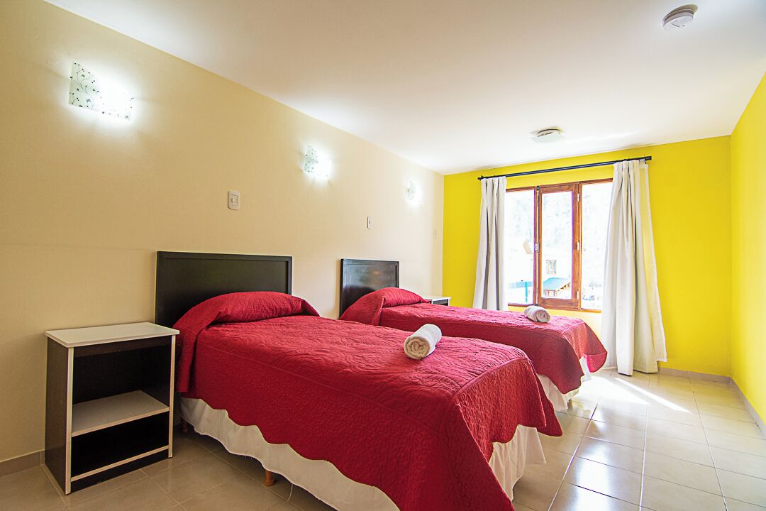 Una habitación con dos camas, una mesita de noche y un ventanal con cortinas.