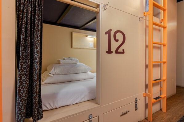 Quarto individual com beliche, o número '12' na parede, uma pequena escrivaninha e uma escada.