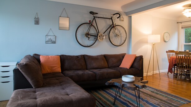 Un salon avec un canapé d’angle marron, un vélo accroché au mur, une table à manger avec des chaises et un lampadaire.