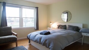 Une chambre à coucher comprenant un lit, une chaise, une table de chevet avec une lampe, un miroir rond et une fenêtre avec des rideaux.
