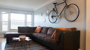Un salon moderne avec un canapé d’angle, un vélo accroché au mur et une grande fenêtre laissant entrer la lumière naturelle.