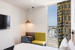 Habitación de hotel con cama, escritorio, silla, lámpara y vista a la ciudad.