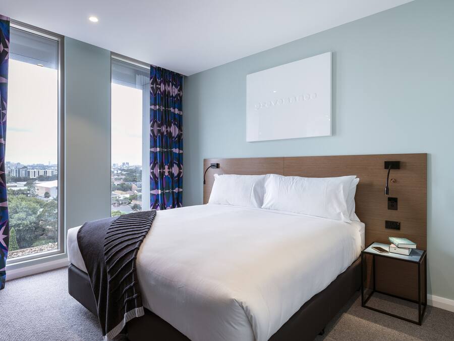 A modern hotel room with a large bed, a nightstand, and a view of the city through large windows.