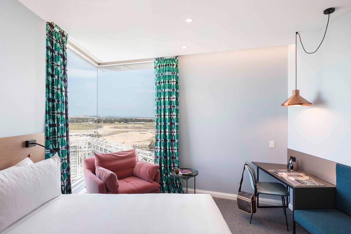 A hotel room with a bed, a pink chair, a small table with a lamp, and a view of a coastal area.