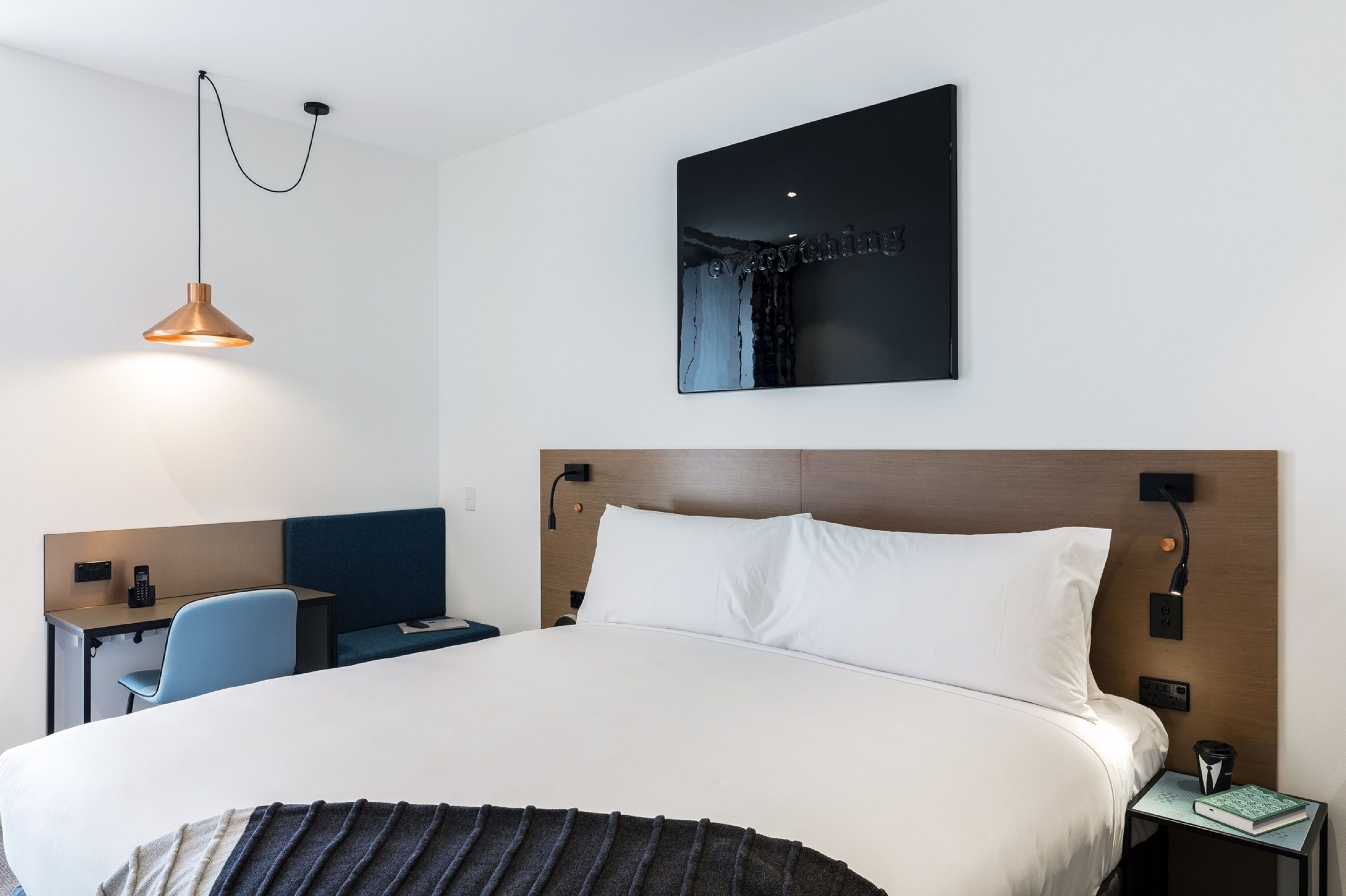 A modern hotel room with a large bed, a desk with a chair, a bedside table with a book, and a wall-mounted artwork.