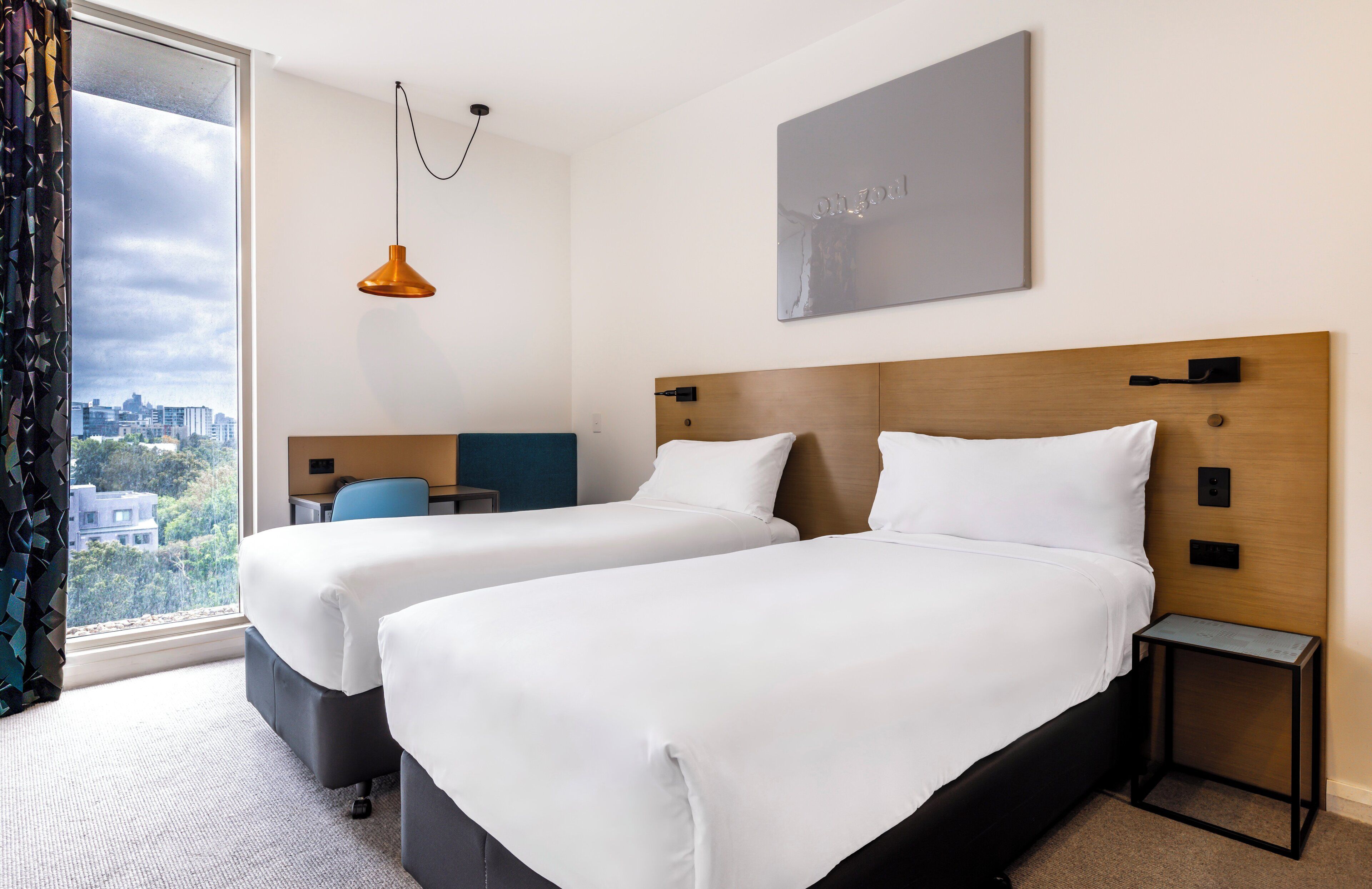 A hotel room with two beds, a city view, a modern pendant light, and a framed artwork on the wall.