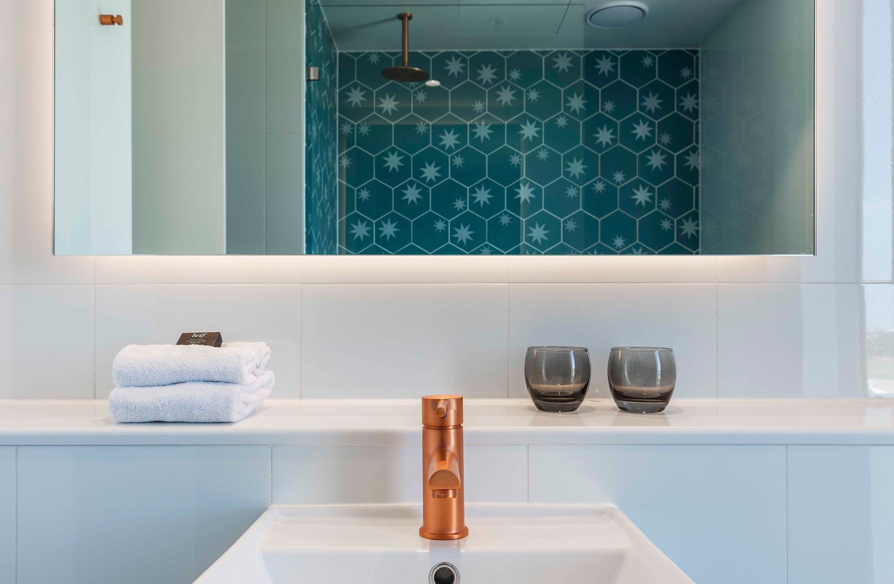 Un baño moderno con un lavamanos blanco, grifo de cobre y un espejo encima. Hay dos toallas apiladas sobre el lavamanos y dos vasos en la encimera.