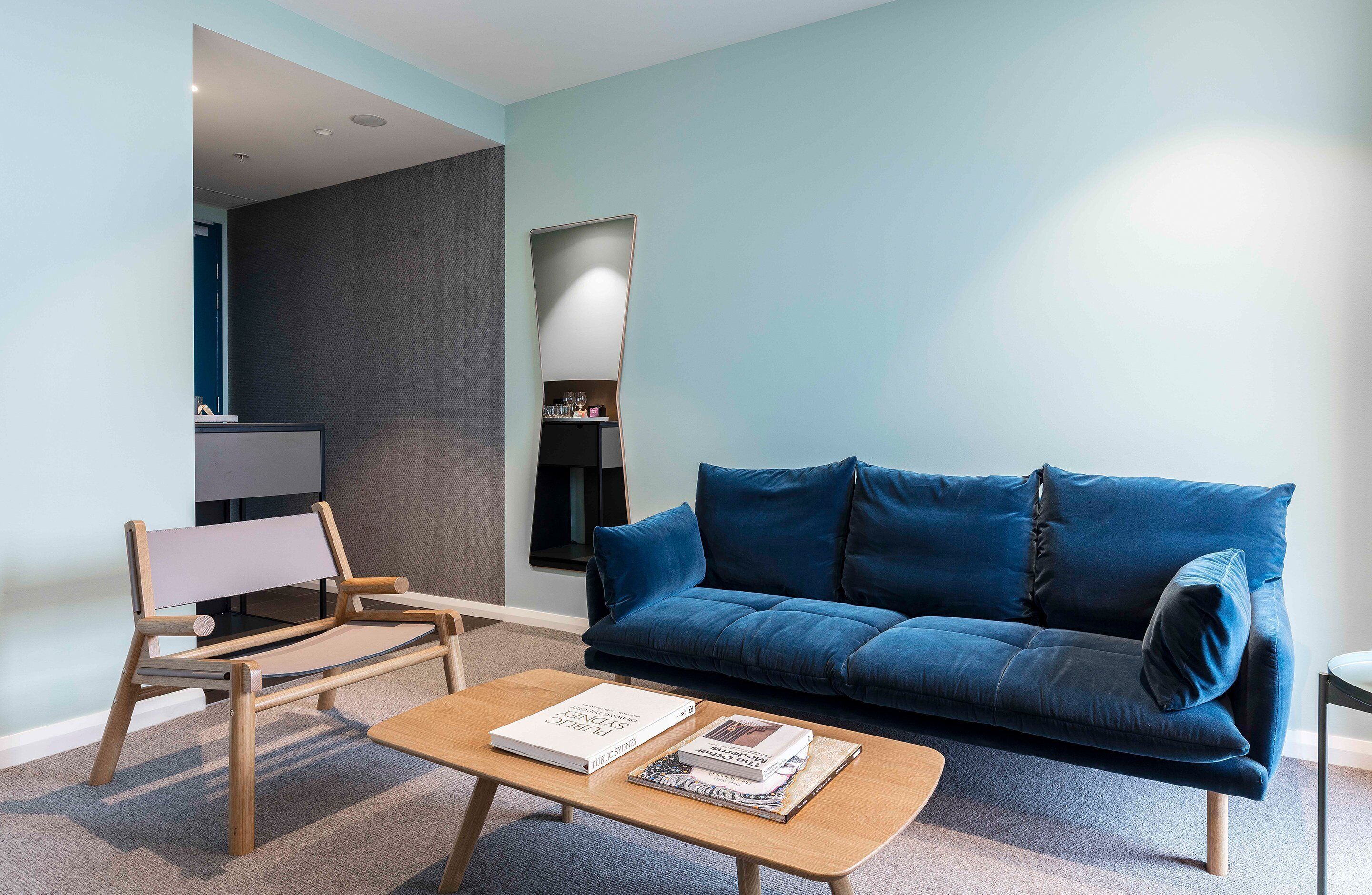A modern living room with a blue sofa, a wooden coffee table with books, a wooden chair, and a dark-colored wall.
