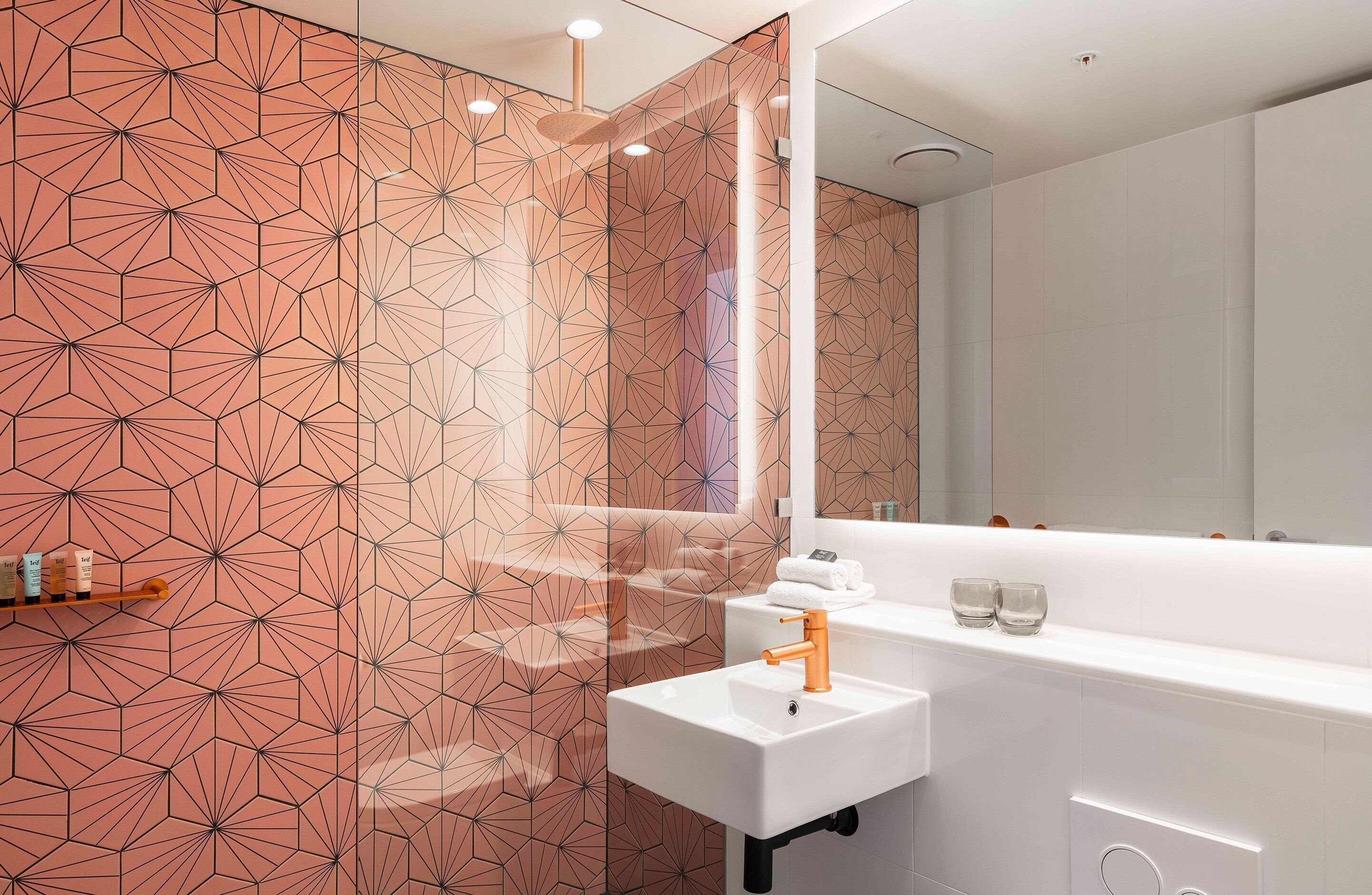 Un baño moderno con pared de diseño geométrico, un lavabo blanco con grifo naranja y un espejo encima.